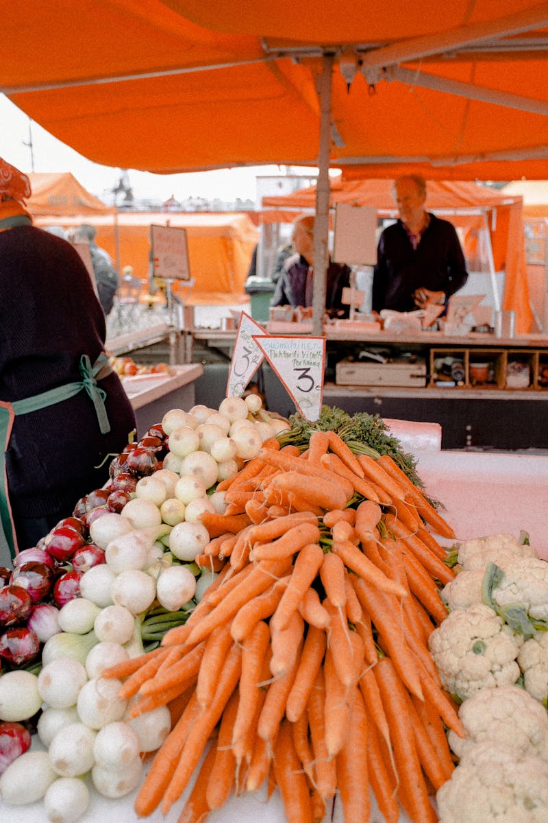 Fresh vegetables displayed at a bustling outdoor market with carrots, onions, and cauliflower.